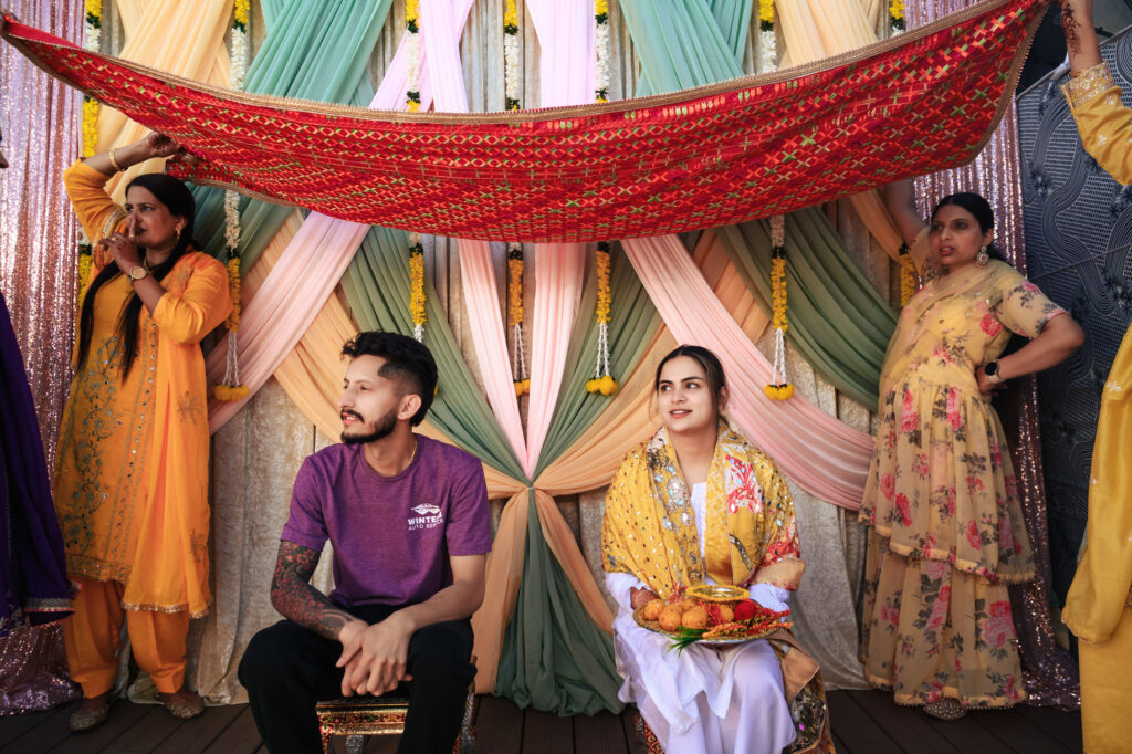 A bride and groom sit under a decorated red cloth during a traditional ceremony. The backdrop features colorful drapes and garlands. Two women in vibrant dresses stand on either side, participating in the event.