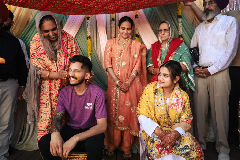 A group of people, dressed in traditional attire, gather happily at a celebration. A man in a purple shirt and a woman in a yellow outfit sit in the center. Colorful drapes and fabric create a festive backdrop.