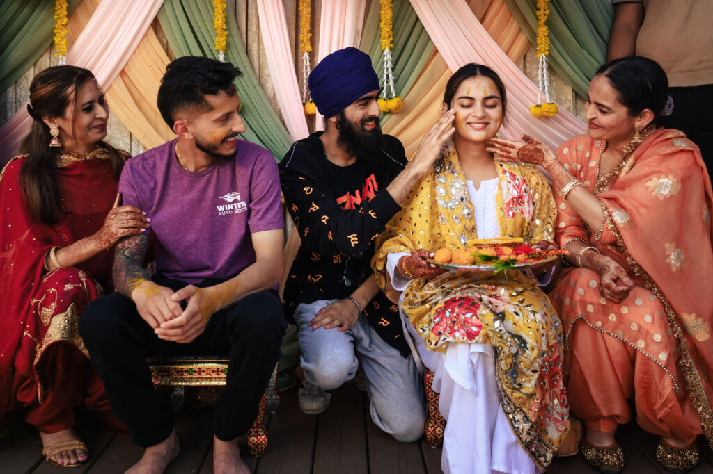 A group of people in colorful traditional attire are celebrating a joyful occasion. In the center, a smiling woman is being affectionately blessed by those around her, holding a decorative tray. Drapes and flowers create a festive backdrop.