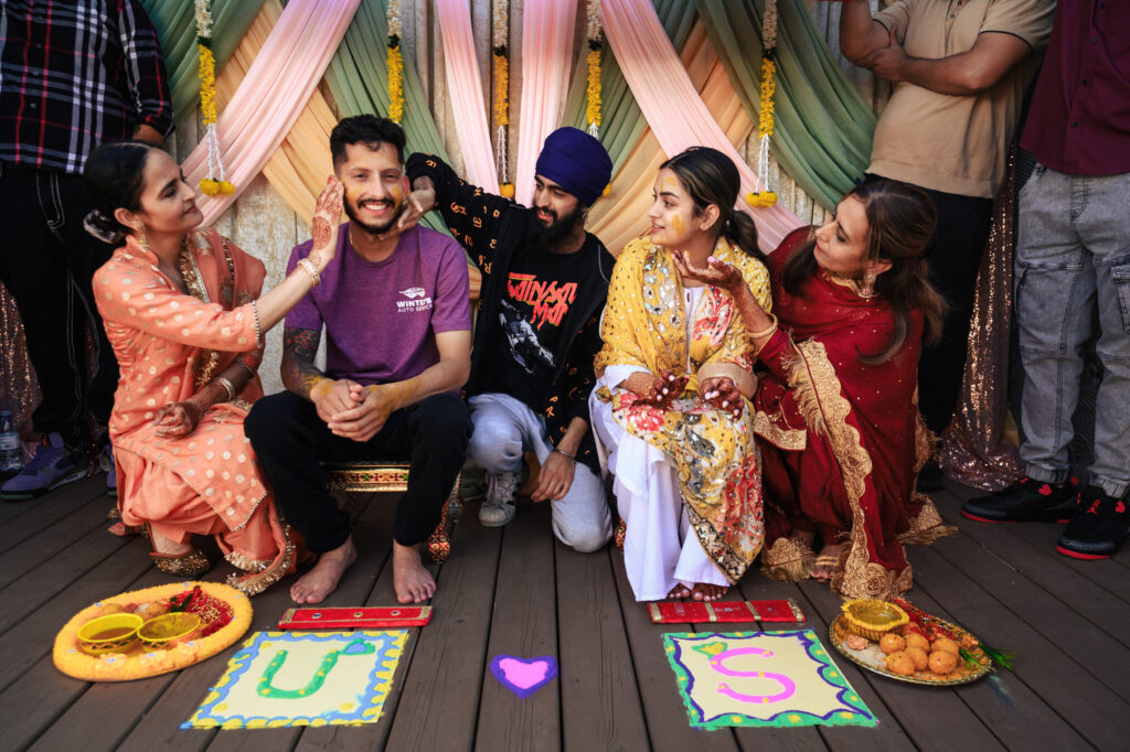A joyful group at a traditional celebration with colorful decorations. Two people are sitting in the center, smiling, while others surround them, playfully touching their faces. Decorative plates with letters "U" and "S" are on the ground.
