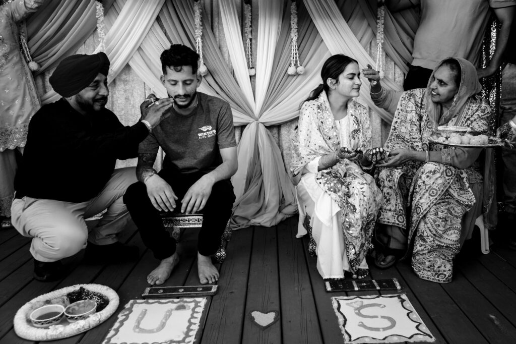 A black and white photo of a traditional ceremony with two people sitting on stools. They are being attended to by older adults. Decorations and a cloth backdrop with initials "U ♥ S" are on the floor.