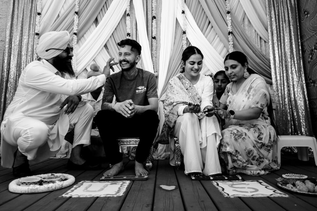 A black-and-white photo of a traditional ceremony with four people. A man in a turban playfully touches the groom, who is smiling. The bride, seated beside him in a decorated outfit, is assisted by a woman. Decorative fabric drapes in the background.