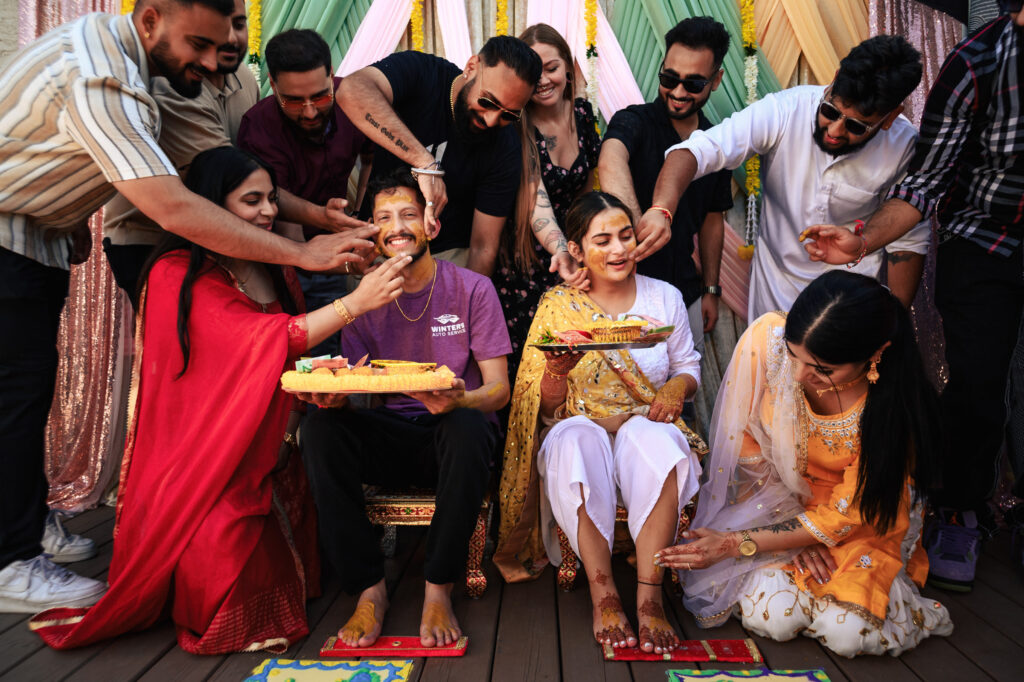 A group of people gather around a seated couple, smiling and playfully applying turmeric paste on them as part of a traditional pre-wedding ceremony. The scene is colorful, with vibrant clothing and festive decorations in the background.