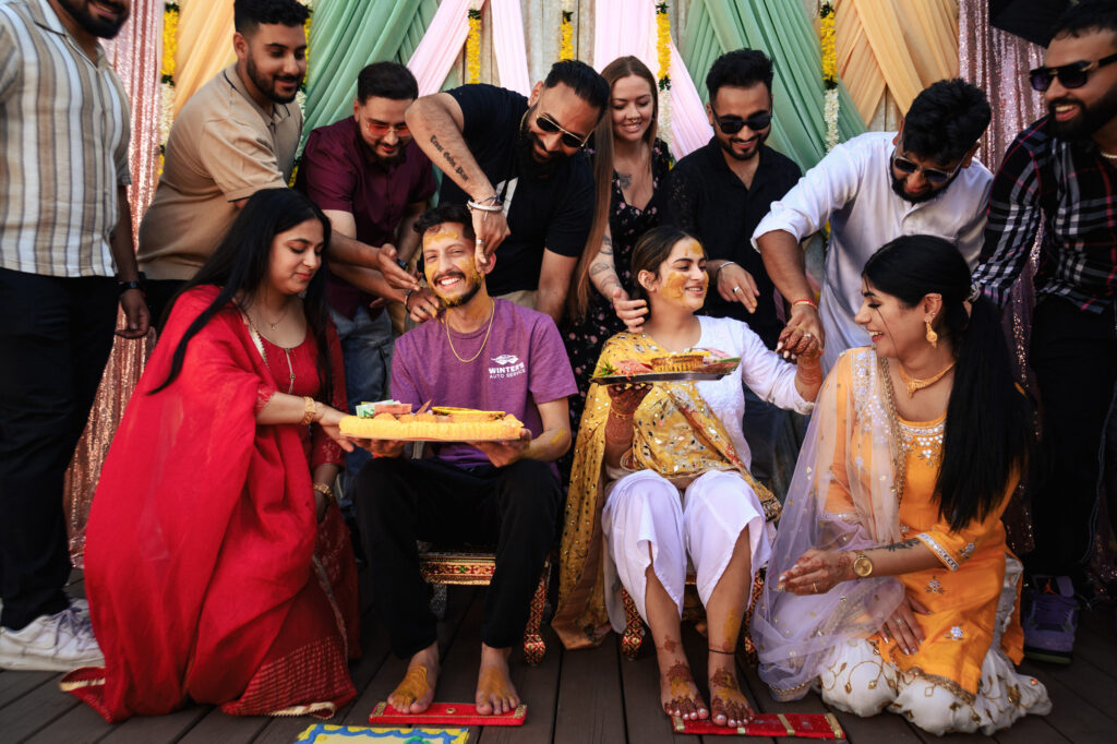 A joyful group of people participate in a traditional ceremony, applying colorful paste to a smiling seated couple. The setting is vibrant, with draped fabrics in the background, creating a festive atmosphere.