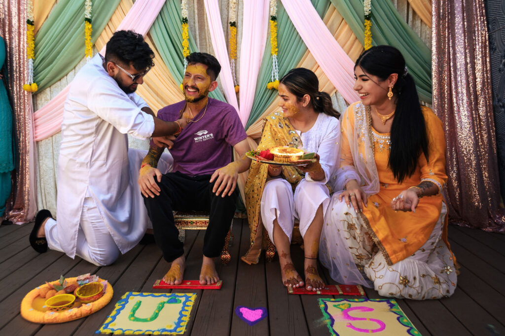 People are celebrating a traditional ceremony with colorful drapes in the background. Two people are having turmeric applied to them, while smiling and sitting between two others. Plates and decorations are on the floor.