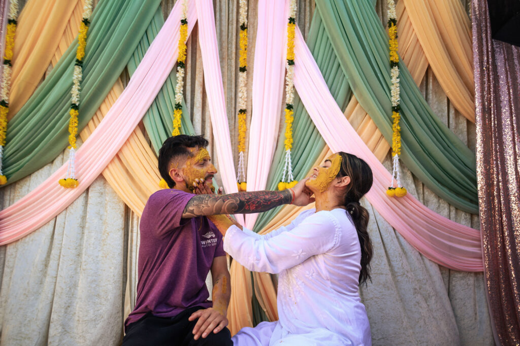 A man and woman sit playfully applying turmeric paste on each other's faces during a traditional ceremony. They are surrounded by colorful drapes and floral garlands. The man wears a purple shirt, and the woman is in a white outfit.