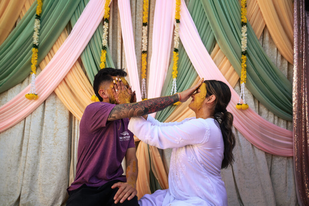 Two people smiling and playfully applying turmeric paste to each other's faces during a traditional ceremony. They are seated in front of colorful draped fabric with floral decorations.