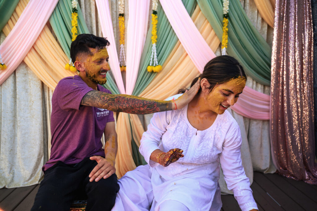 A joyful couple participates in a traditional pre-wedding ceremony, smiling as they apply turmeric paste on each other. They sit in front of a decorative backdrop with pastel-colored drapes and marigold flowers.