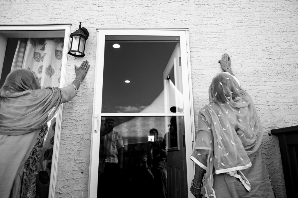 Two women in traditional clothing touch the outside walls of a house near the door, gazing up. Their gestures seem to convey reverence or blessing. Through the glass, reflections of people are visible inside. The image is in black and white.