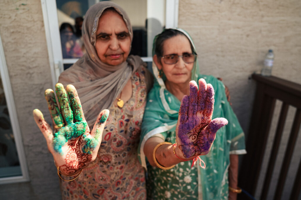 Two older women wearing traditional South Asian attire show their hands covered in colorful powder and intricate henna designs, standing outdoors. The woman on the left wears beige, while the one on the right wears green.