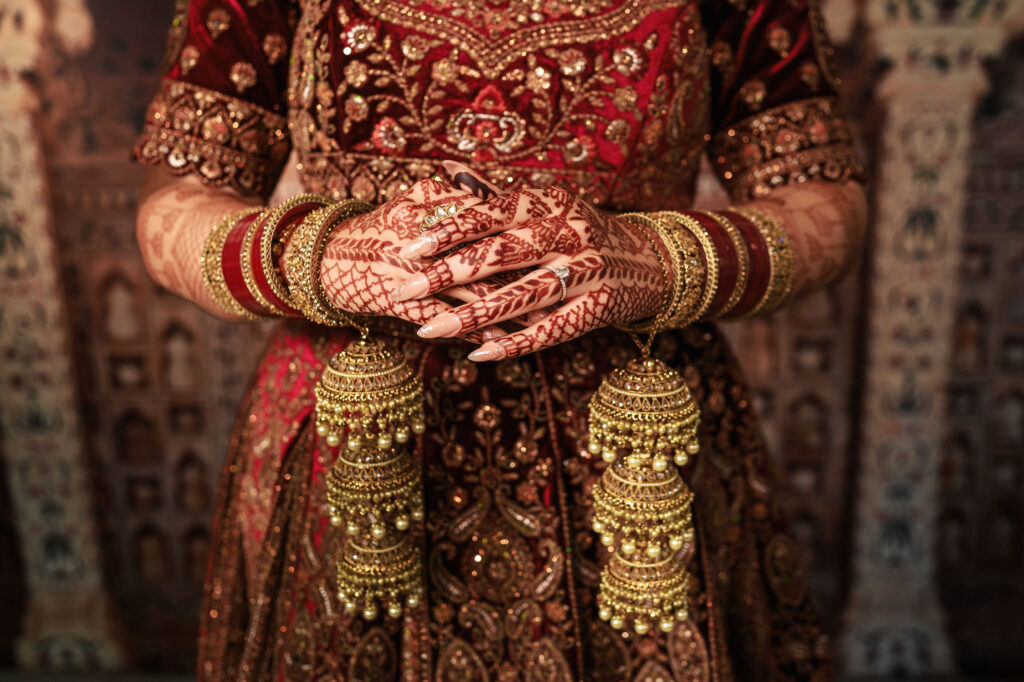 A person wearing an ornate red and gold outfit, adorned with intricate henna on their hands. They are also wearing multiple gold bangles and holding large, decorative tassels. The background features detailed, architectural patterns.