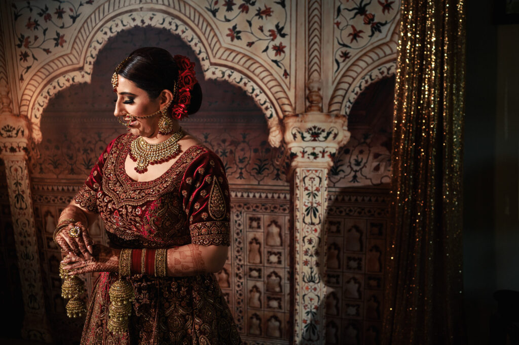 A woman in traditional Indian bridal attire, adorned with intricate jewelry and henna, stands in front of a richly decorated backdrop. She gazes to the side, with her hands elegantly positioned, highlighting a detailed outfit.