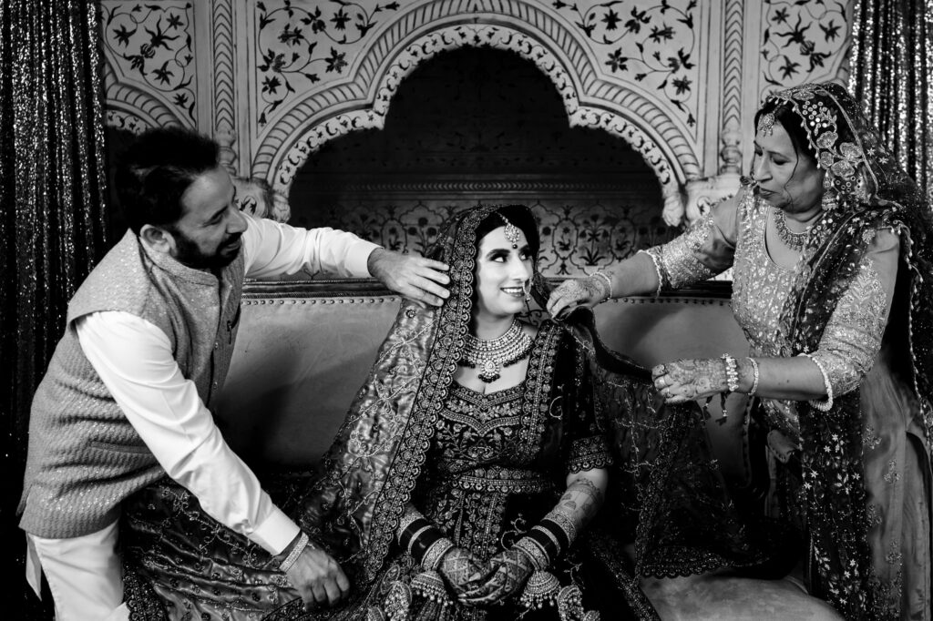 A bride in traditional attire is seated on a sofa, smiling as a man and woman, possibly her parents, lovingly adjust her veil. The background is ornately decorated, capturing a festive and joyful atmosphere.