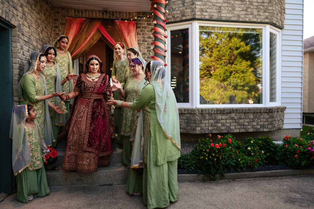 A bride in a traditional burgundy and gold outfit stands surrounded by women in matching green dresses and headscarves. They are near a brick building with decorative flowers, and a tree is reflected in a window nearby.