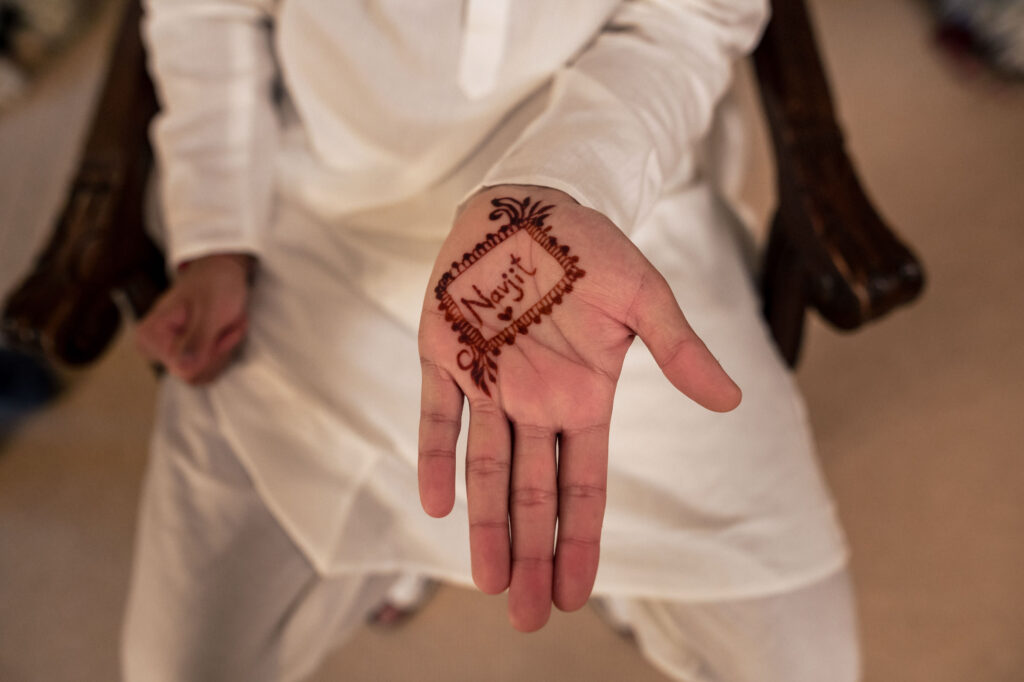 A person in white traditional attire is showing their open hand with intricate henna designs, including the word "Nazij." The background is softly blurred.