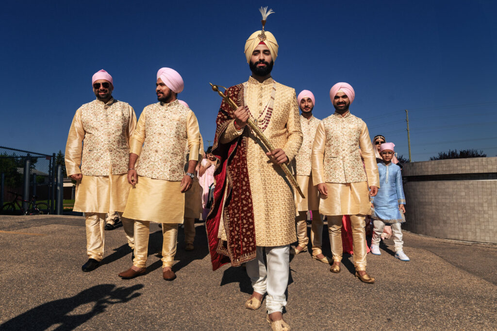 A groom in traditional Indian attire, holding a staff, walks confidently at the front. He is accompanied by five groomsmen wearing similar outfits and pink turbans, under a bright blue sky. A young child in a light blue outfit follows them.