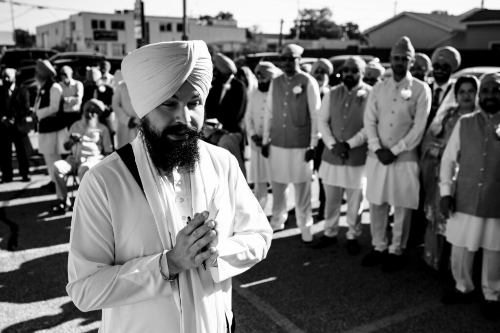 A man wearing a turban and traditional attire stands with hands clasped in front of a group of similarly dressed individuals. They're outdoors, possibly during a ceremony or celebration. The image is in black and white.