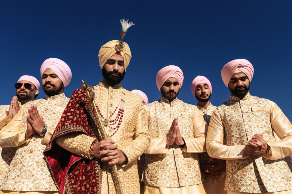 A groom in traditional Indian attire, holding a ceremonial sword, poses with five groomsmen wearing matching pale pink turbans and floral-patterned sherwanis. They stand against a clear blue sky, with the groomsmen holding their hands in a respectful gesture.