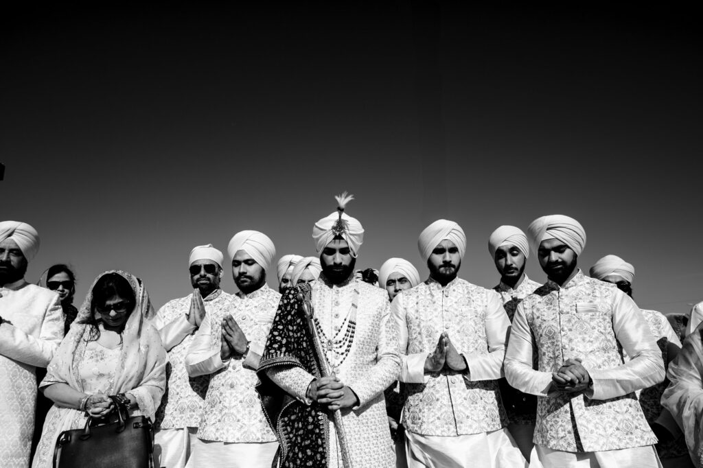 A group of men dressed in traditional Indian attire, including turbans and ornate sherwanis, stand together. The central figure holds a staff, while others have their hands in a prayer position, in what appears to be a wedding ceremony.