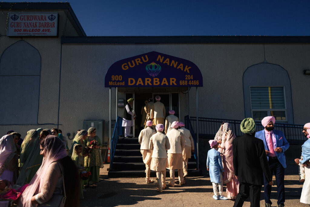 A group of people dressed in traditional attire enter a building labeled "Guru Nanak Darbar." The entrance is decorated, and the sign includes the address and phone number. The sky is clear and blue, and some people are conversing outside.