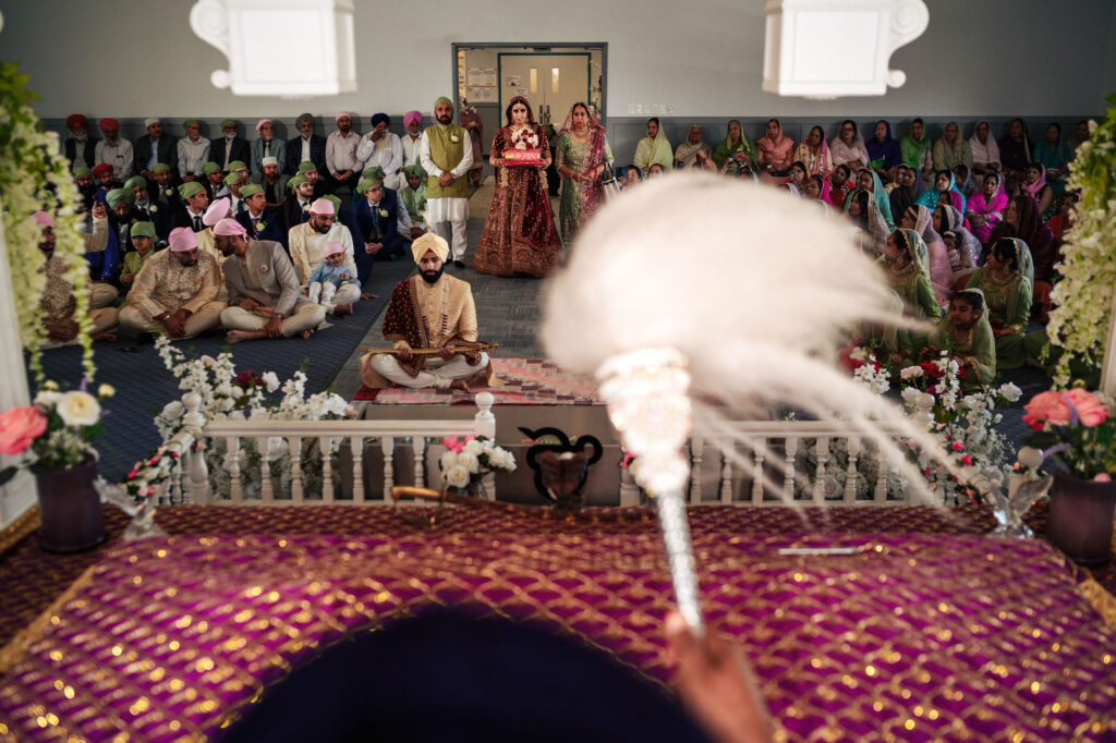 A Sikh wedding ceremony inside a gurdwara. The couple sits at the front near the Guru Granth Sahib, with guests seated behind them. The priest holds the chaur sahib. The room is adorned with flowers.