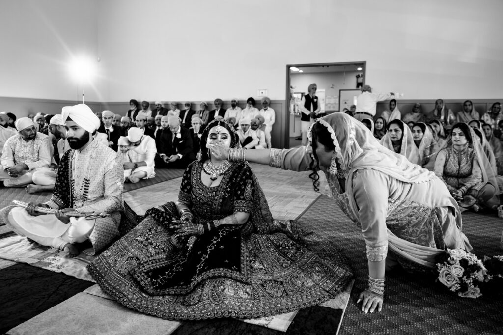 A black-and-white photo of a Sikh wedding ceremony. The bride and groom sit on the floor wearing traditional attire. A woman adjusts the bride's jewelry. Guests are seated in the background, observing the ritual.