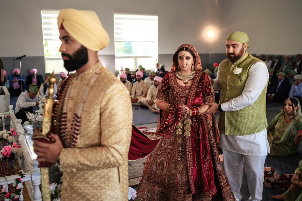 A bride and groom in traditional attire participate in a wedding ceremony at a Sikh temple. The bride wears a red lehenga, and the groom is in a cream outfit with a turban. Guests are seated in the background.