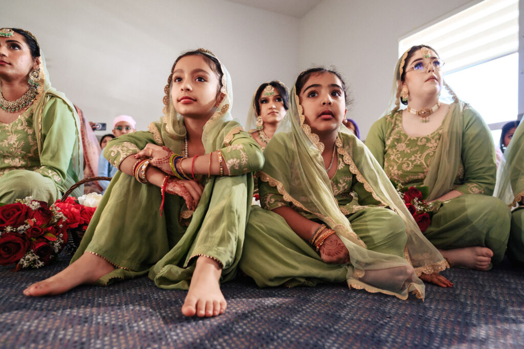 Two young girls in green traditional attire sit on a carpeted floor, surrounded by people in similar outfits. One girl sits cross-legged, while the other sits with legs outstretched, both focused on something ahead.