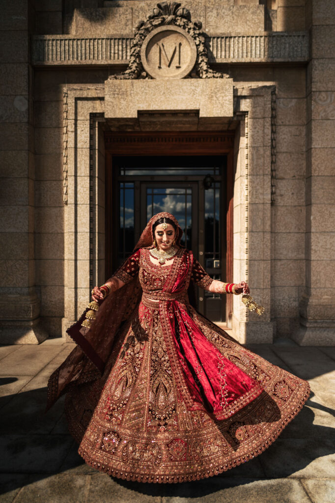 A woman in an ornate red and gold traditional dress poses gracefully in front of a grand stone building with a decorative entrance. She is adorned with jewelry and the sunlight highlights the intricate details of her attire.