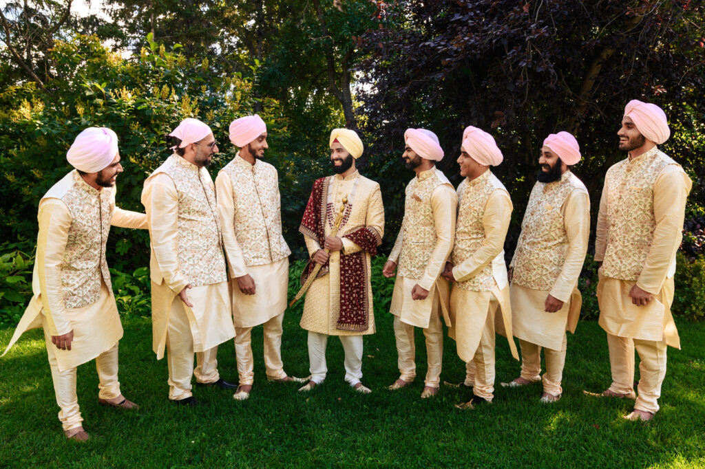 A groom in ornate traditional attire stands smiling with seven groomsmen wearing cream-colored sherwanis and pink turbans. They are outdoors on vibrant green grass, surrounded by lush trees and bushes.