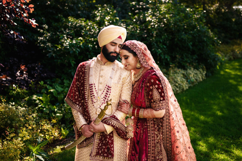 A couple dressed in traditional South Asian attire stand closely in a lush garden. The man wears a cream and maroon sherwani with a turban, holding a ceremonial sword. The woman is adorned in a heavily embroidered maroon and gold lehenga with jewelry.