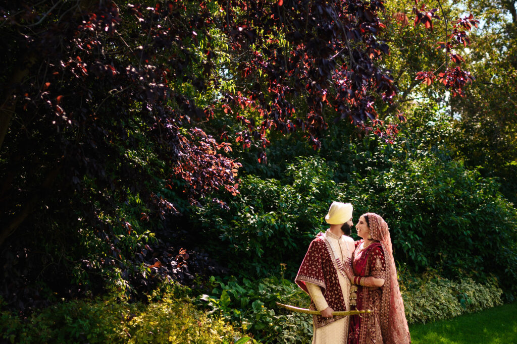 A couple dressed in traditional South Asian wedding attire stands closely in a lush garden. The bride wears a red outfit with intricate designs, and the groom wears a cream and red ensemble. Sunlight filters through the trees, casting a warm glow.