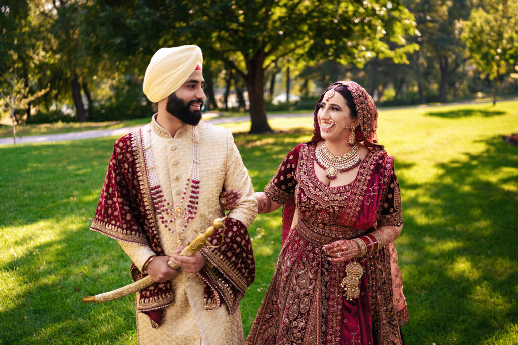 A couple dressed in elegant traditional South Asian attire stands in a sunny park. The man wears a cream-colored sherwani and turban, holding a sword. The woman is in a red and gold lehenga, adorned with jewelry. They smile at each other.