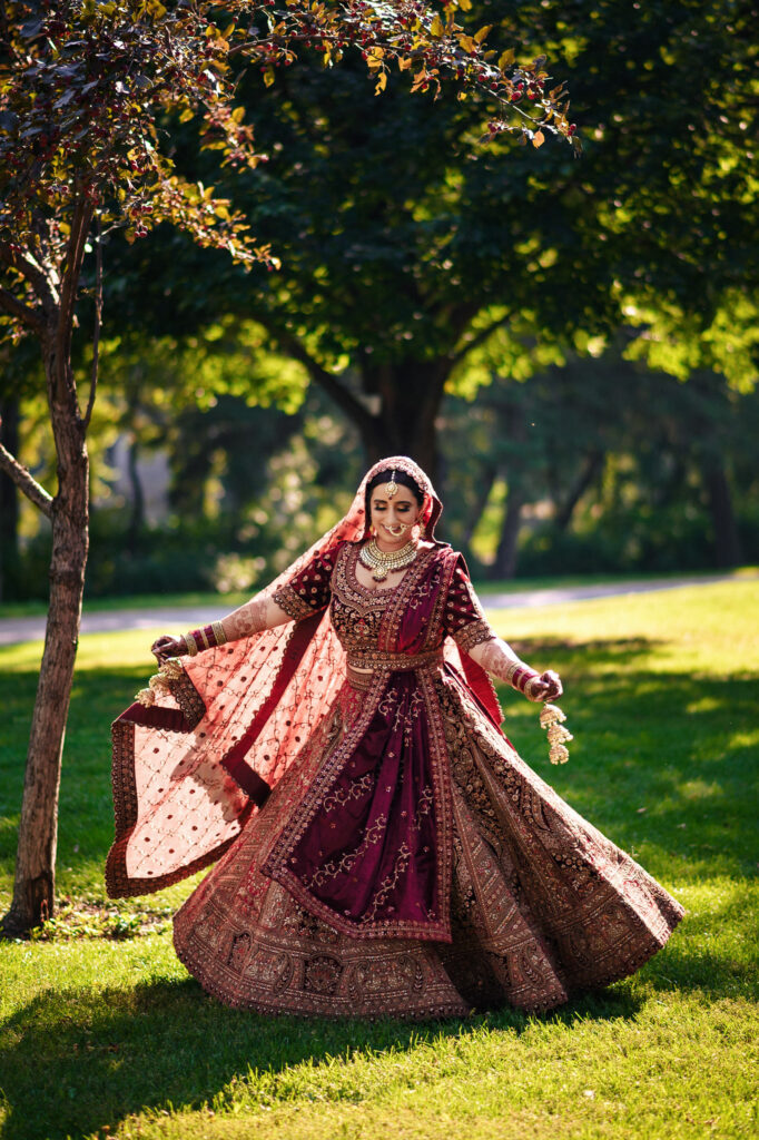 A woman twirls in a burgundy and gold wedding dress adorned with intricate embroidery. She wears traditional jewelry and a veil, surrounded by lush greenery and sunlight filtering through the trees.