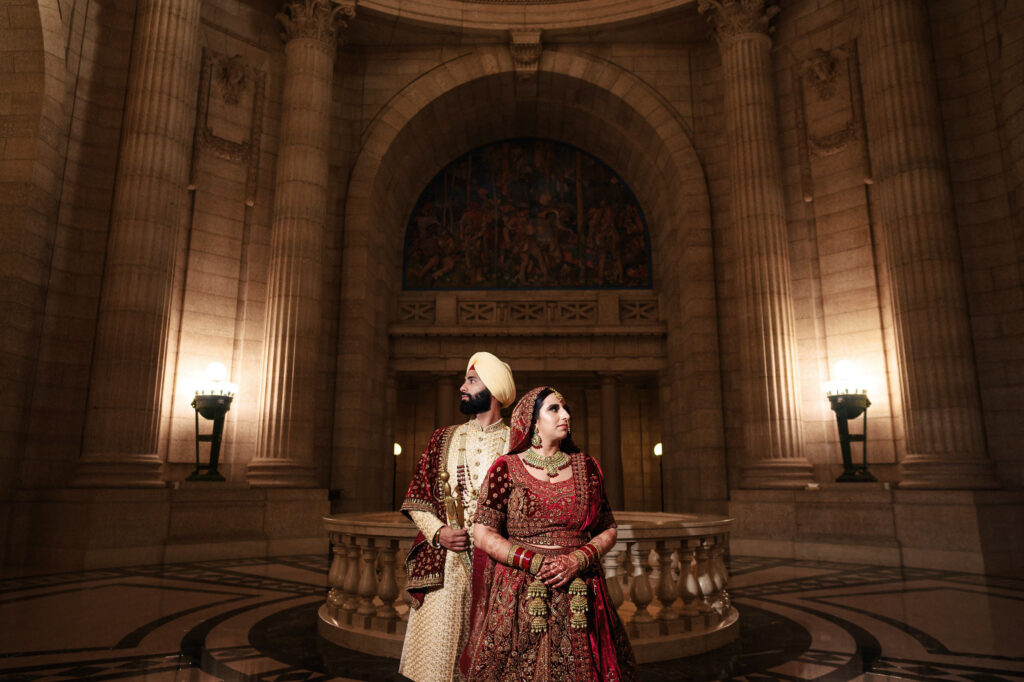 A couple stands in an ornate hall with tall stone columns and intricate wall carvings. The man wears a cream sherwani and turban, while the woman is in a red and gold lehenga. They look in opposite directions, exuding elegance and tradition.