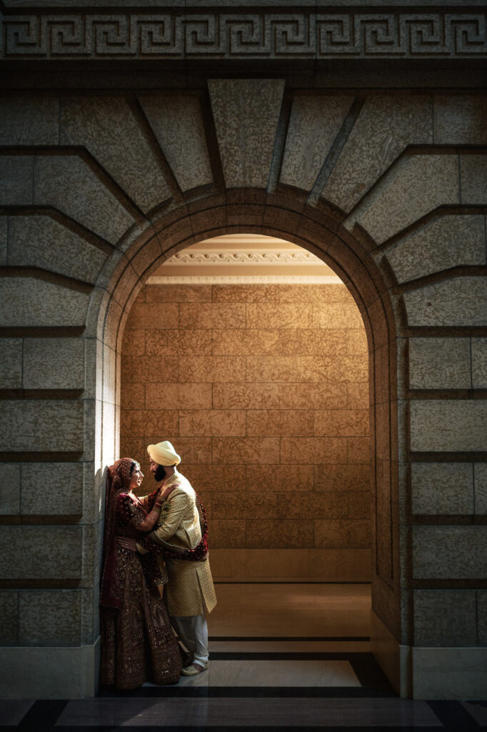 A couple dressed in traditional Indian wedding attire shares a tender moment under an illuminated stone archway. The bride is in a red lehenga, and the groom wears a cream sherwani with a turban, surrounded by intricate stone patterns.