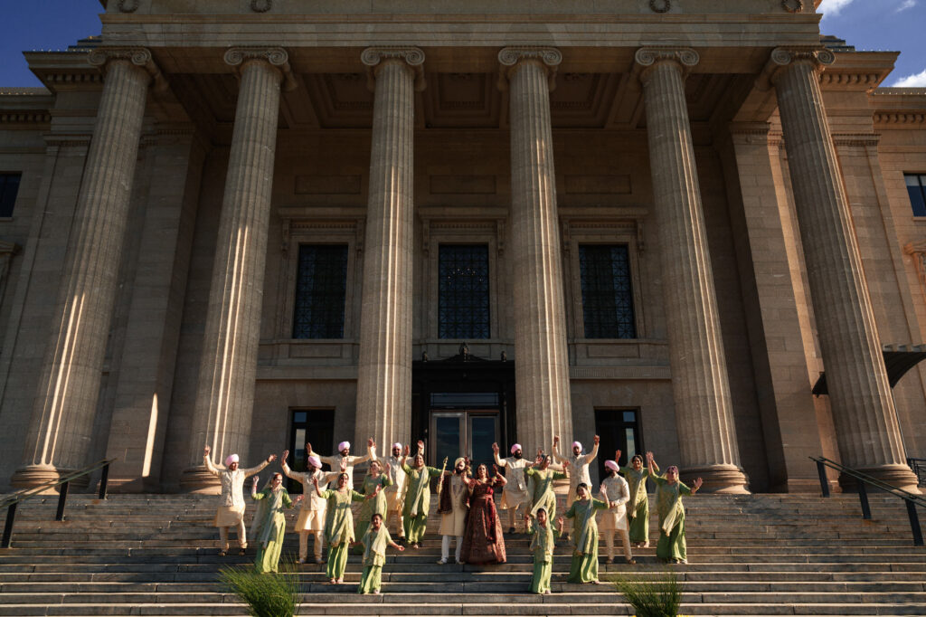 A group of people in traditional attire celebrate on the steps of a grand building with tall columns. The building's entrance is framed by large, decorative columns under a clear blue sky.