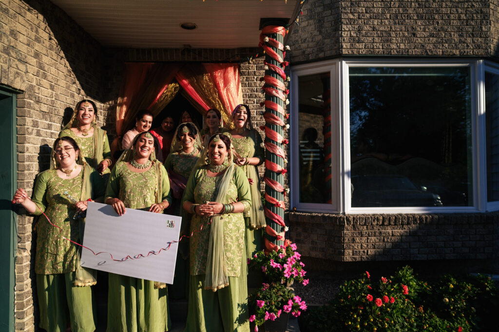A group of women in matching green traditional attire stand on a porch, holding a decorated sign with a red ribbon. The porch is adorned with colorful drapes and flowers, and the setting sun casts warm light on the scene.