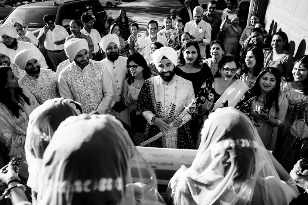 A joyous wedding scene shows a groom holding a ceremonial sword, surrounded by smiling guests dressed in traditional attire. The black and white photo captures the celebratory atmosphere of a cultural gathering.