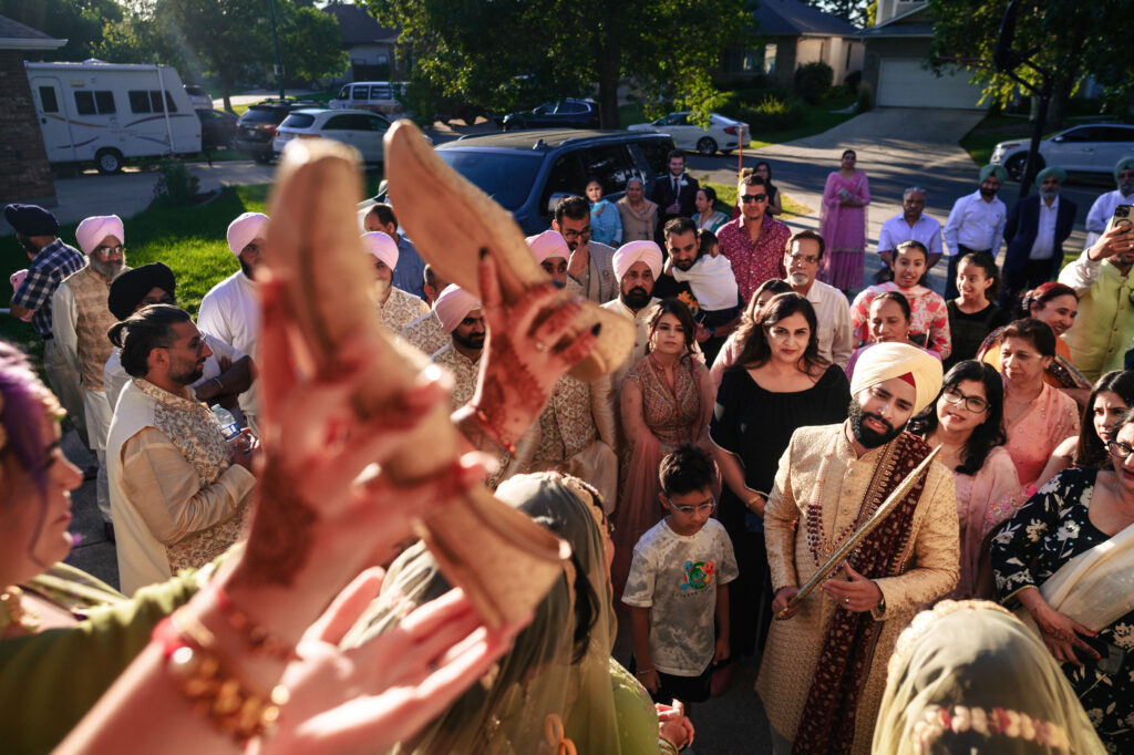 A festive gathering of people in traditional attire, including turbans and dresses, captures a joyful outdoor celebration. A pair of shoes is humorously held up in the foreground, with a beaming crowd in the background under a sunny sky.