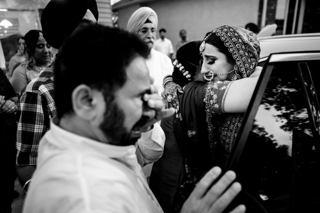 A bride in traditional attire hugs a woman, surrounded by a group of people. A man in the foreground appears emotional, covering his face with his hand. The scene conveys a poignant farewell moment, possibly at a wedding.