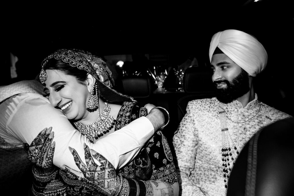 A bride and groom, dressed in traditional Indian wedding attire, sit in a car. The bride, adorned with intricate jewelry, is smiling as she hugs someone who is partly visible. The groom watches with a gentle smile. The image is in black and white.