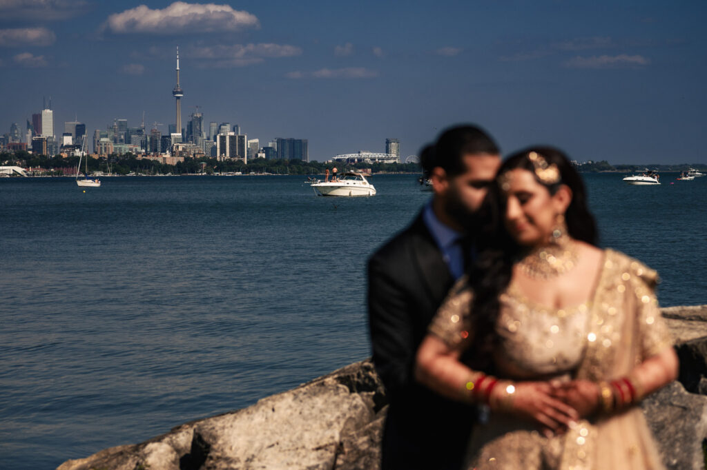 A couple in traditional attire stands on a rocky shore with a blurred Toronto skyline, including the CN Tower, in the background. The sunlit scene captures a serene lake view with boats sailing under a blue sky.