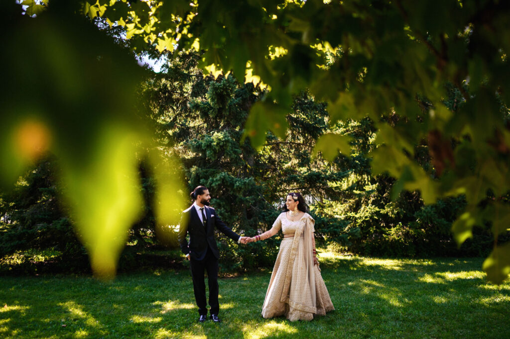 A couple holding hands and walking on a grassy lawn under the shade of trees. The woman is wearing a flowing beige gown, and the man is in a dark suit. Sunlight filters through the leaves, creating a dappled effect on the ground.