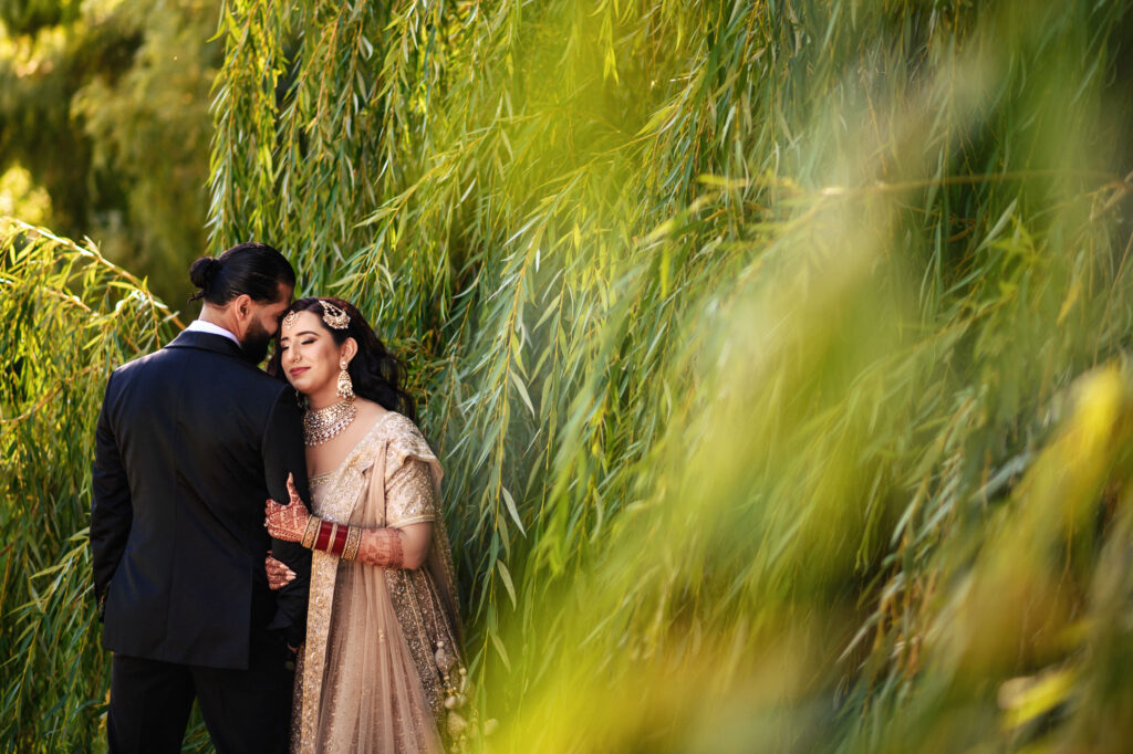 A couple stands together, with the woman leaning her head on the man's shoulder. She is wearing a traditional embellished dress, jewelry, and bangles. The setting is lush and green, with tall, leafy branches surrounding them.
