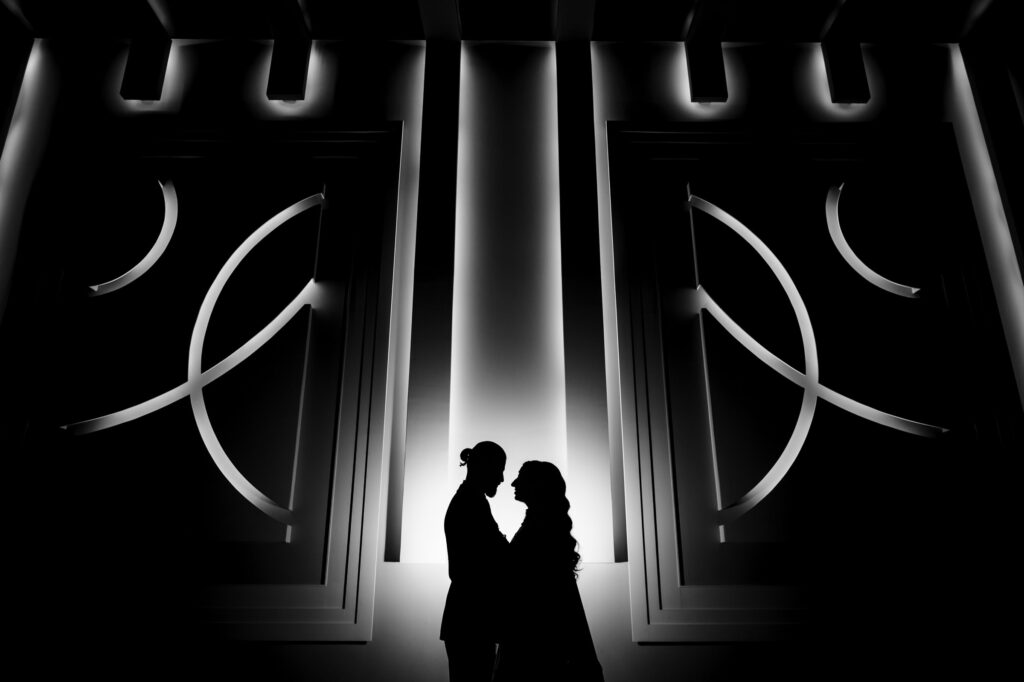 Two people in silhouette stand facing each other against a backdrop of large illuminated geometric designs. The black and white composition creates a dramatic and intimate atmosphere.