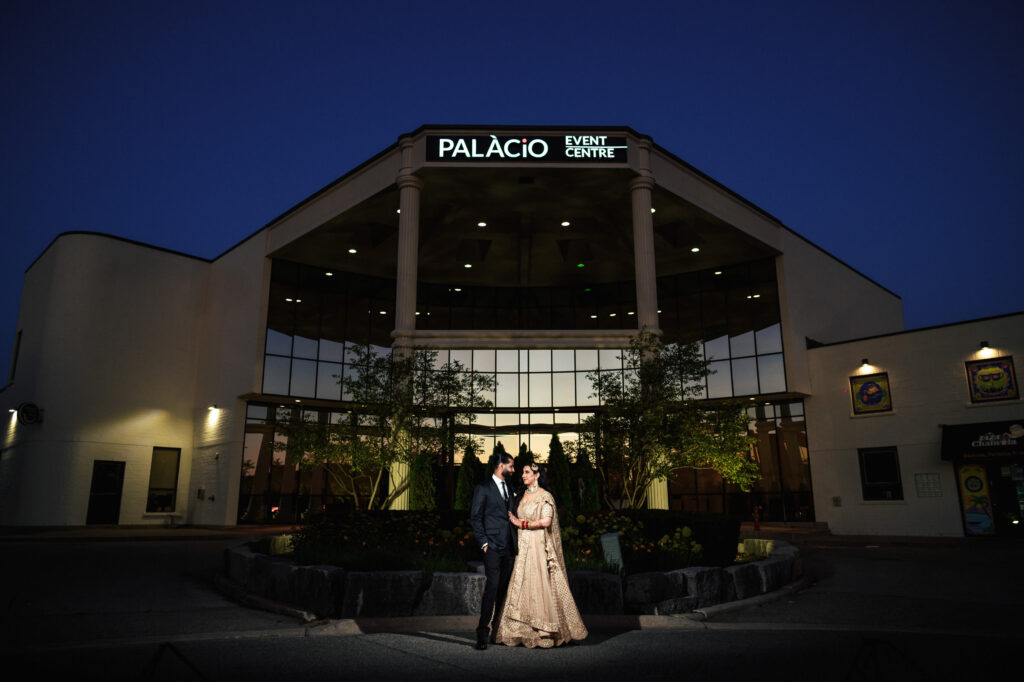 A couple dressed in formal attire stand outside the Palacio Event Centre at night. The building is brightly lit, showcasing its glass facade and towering columns. Trees and plants can be seen in the foreground.