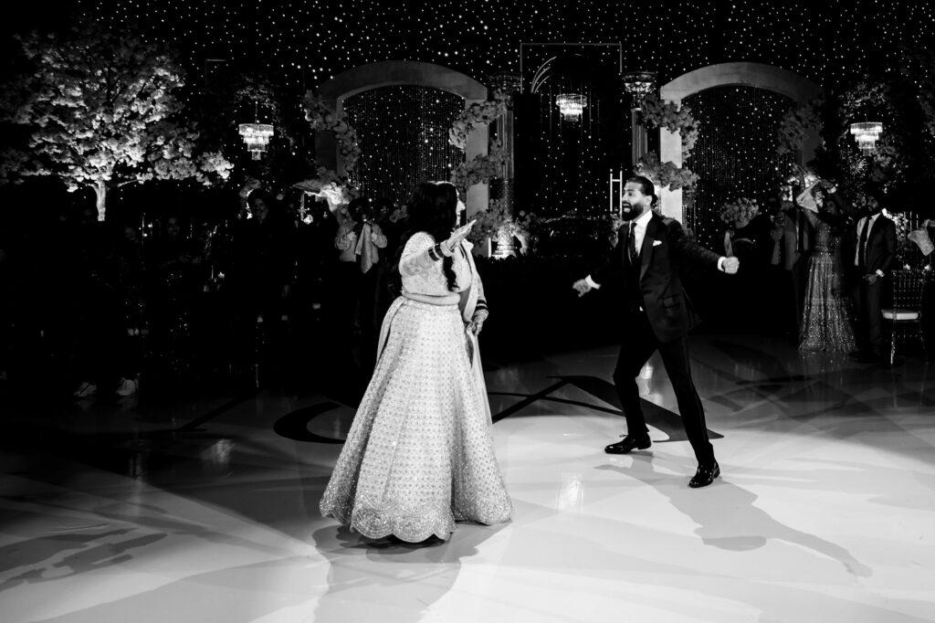 A couple dances joyfully at a formal event. The woman wears a traditional gown, and the man is in a suit. The venue is elegantly decorated with twinkling lights and arches, and several guests are visible in the background.