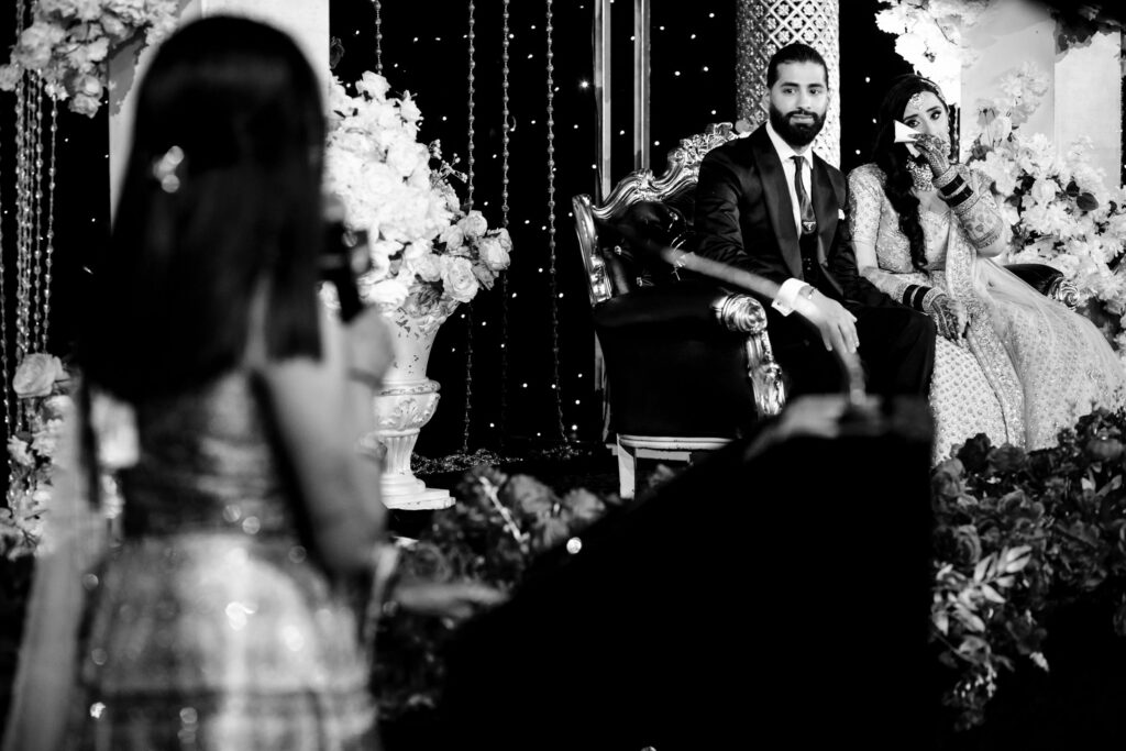 A black and white photo of a couple sitting on a decorated stage, dressed in formal attire. The woman wears a traditional dress and jewelry. In the foreground, a blurred person appears to be speaking or singing into a microphone. Floral arrangements adorn the stage.