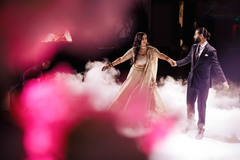 A couple dances on a fog-covered floor, holding hands and smiling. The woman wears a traditional dress, while the man is in a dark suit. Pink and red flower petals blur in the foreground, adding vibrant color to the scene.